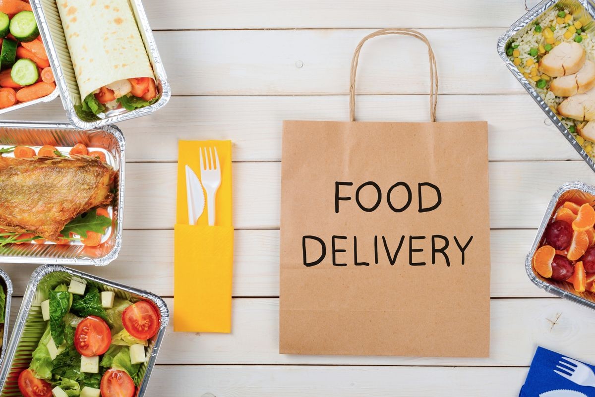Fish, a vegetable roll, tomato and lettuce salad, plastic flatware and a paper bag with Food Delivery sign, close-up. Rice with chicken, fruit, wooden surface. Dinner for busy people.